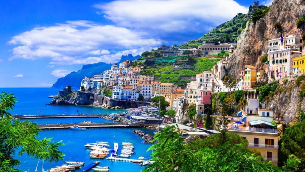 Panoramic view of the colorful buildings of Positano cascading down the hillside to a beach dotted with umbrellas, with boats dotting the azure sea."