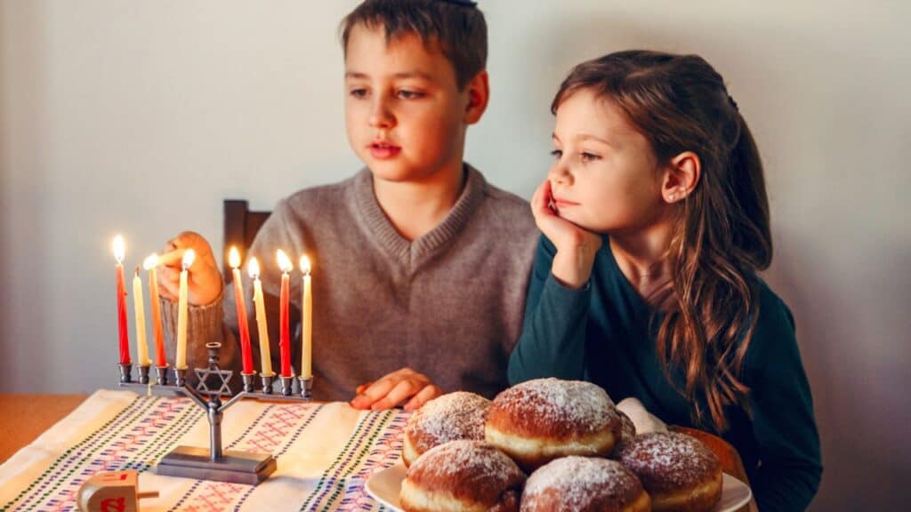 Brother and sister siblings lighting candles on menorah for Jewish Hanukkah holiday at home. Children celebrating Chanukah festival of lights. Dreidel and Sufganiyot donuts in plate on table.