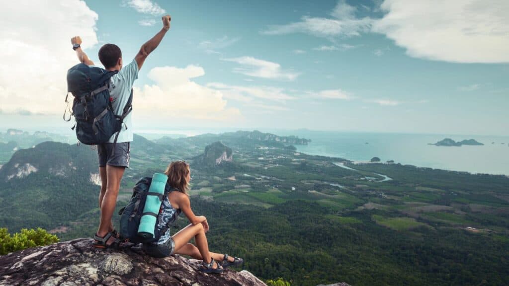 Two hikers at the top of the mountain. One has their arms raised above their head triumphantly.