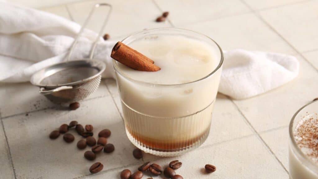 Glass of White Russian cocktail with cinnamon and coffee beans on white tile table