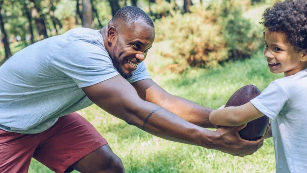 Cheerful african american man playing football with adorable son in park