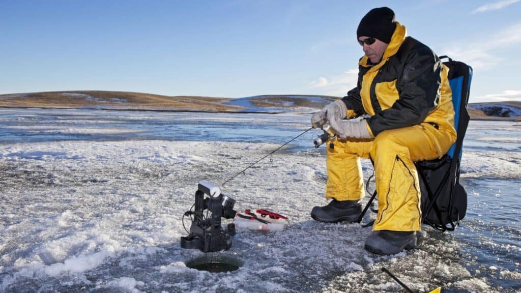 A guy in a yellow snowsuit sits next to a hole in the ice on a lake fishing