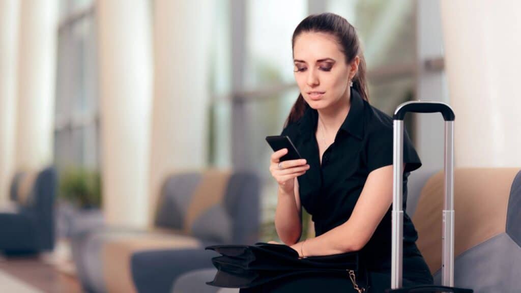 Young woman with a suitcase sitting at the airport on her phone.