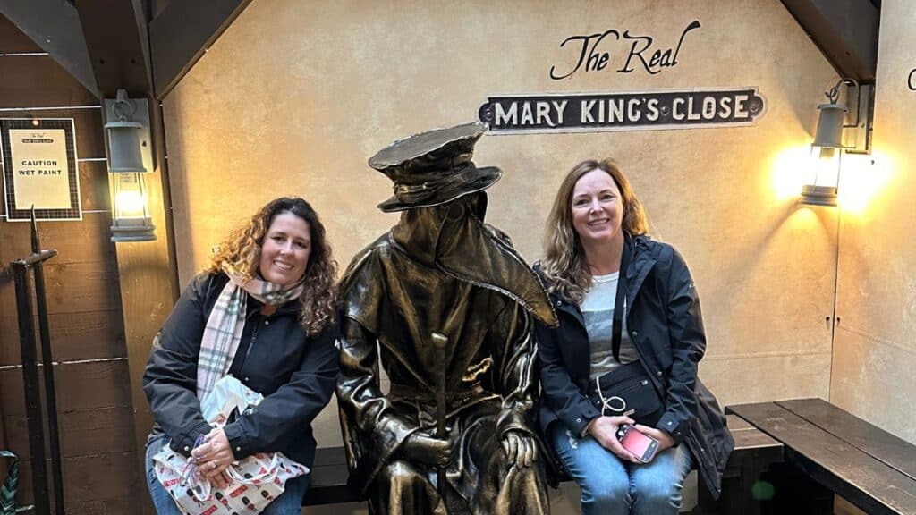 Two women sitting with a plague doctor statue outside the Real Mary King's Close in Edinburgh Scotland